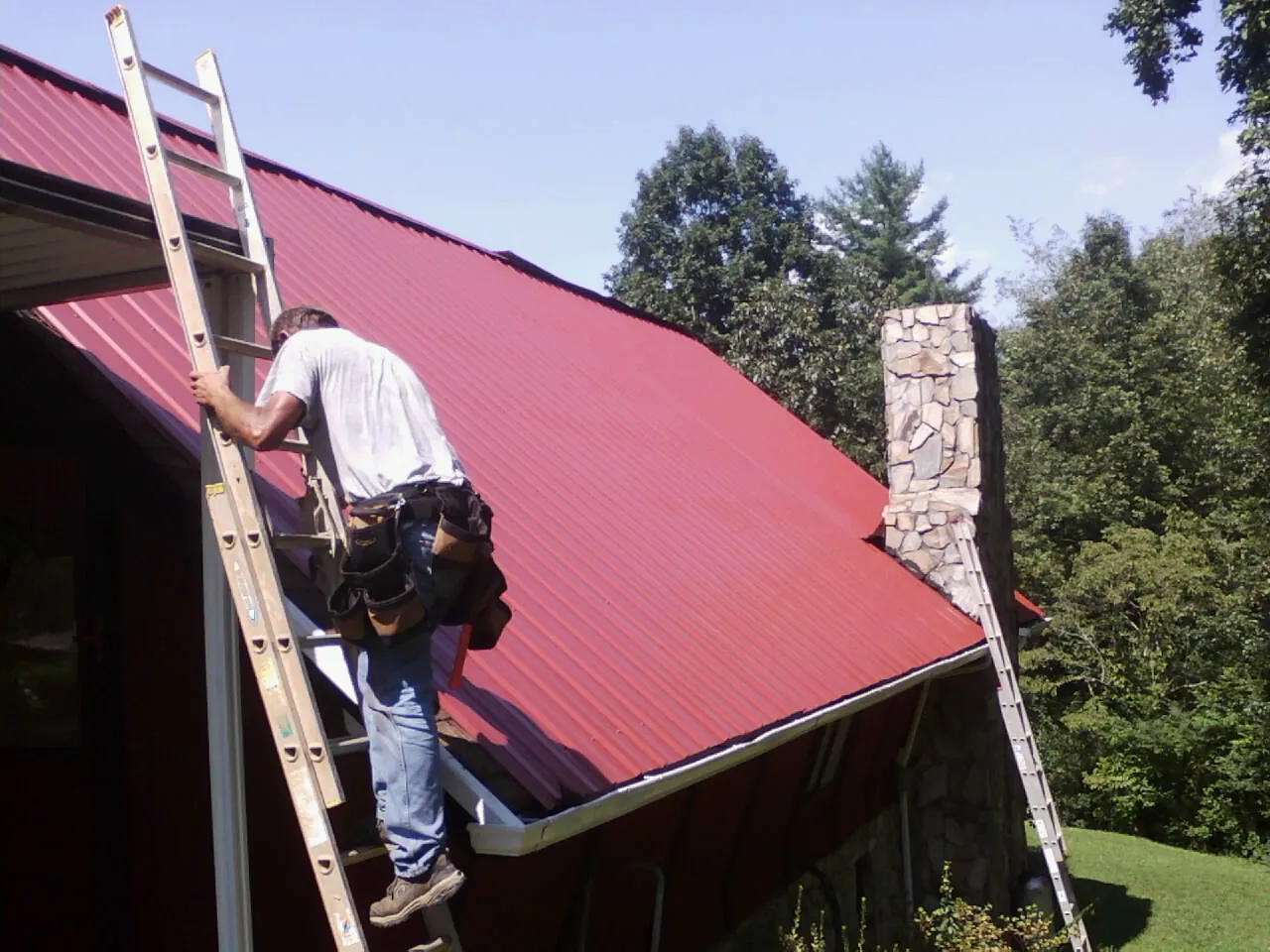 Roofer on a ladder beside a red metal roof and stone chimney — Soffit & Fascia Repair by Roofing Brothers in La Grange